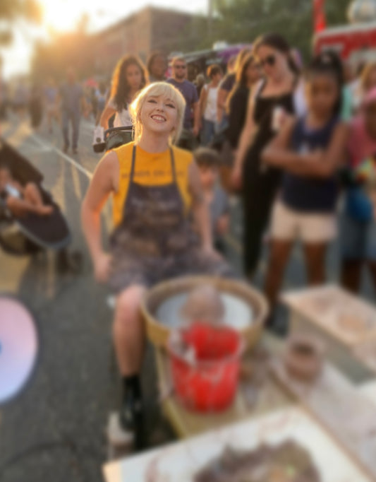 Meghan sitting at a pottery wheel on the street with audience’s faces blurred.